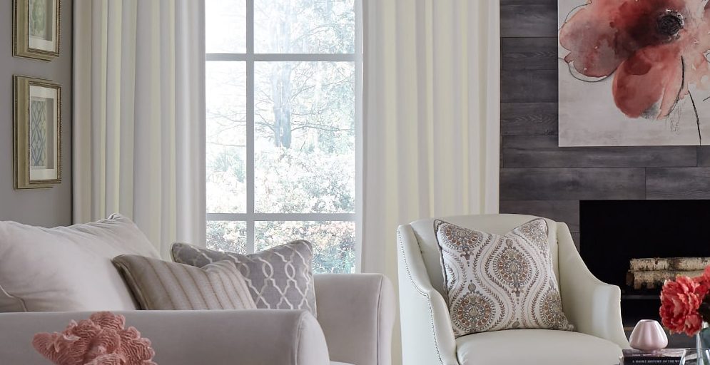 Large window featuring drapes partially drawn in front of a table with a plant on top.